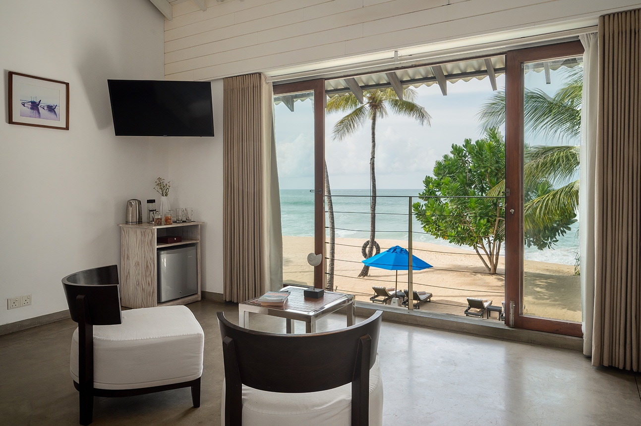 An upstairs bedroom's sitting corner with 2 armchairs, and glass doors to a balcony over the beach, a blue umbrella and the sea
