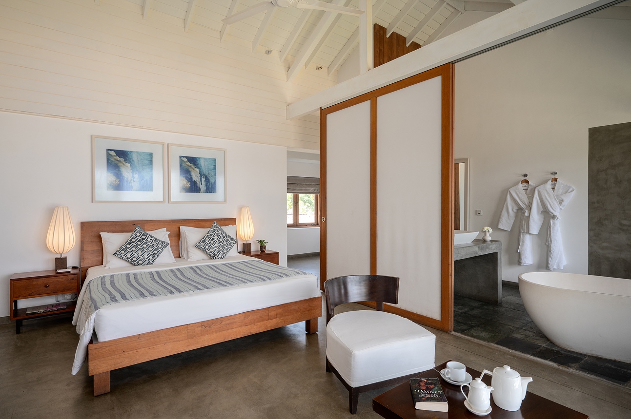 A bedroom with a timber bed, an armchair and a freestanding bathtub behind an open sliding panel