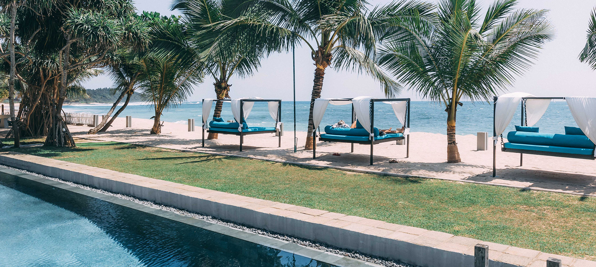 Curtained daybeds with blue cushions on the sand under palms, with the pool and lawn in the foreground
